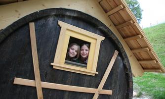 Two girls are happily looking out of a small window of a rustic wooden house. The house has a round shape and a cozy atmosphere.