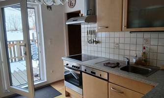 A modern kitchen with light wooden furniture and an open door leading to an outdoor area. The space is well-lit and clean.
