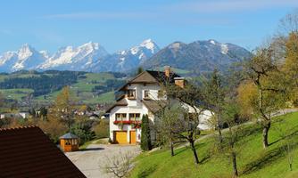 A picturesque house amidst green meadows with snow-capped mountains in the background. The landscape is sunny and inviting.