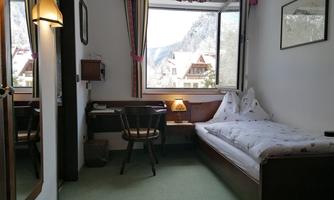A simple room with a single bed, a desk, and a chair. Large windows offer a view of the mountains.