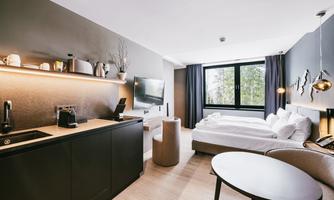 A modern hotel room with a kitchenette, a double bed, and a cozy seating area. Large windows let in plenty of daylight and offer a view of greenery.