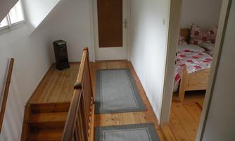 A bright hallway with a wooden staircase and carpets. On the left is a room with a bed and a blanket in red and white.