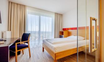 A modern hotel room with a large window that lets in plenty of light. There is a double bed, a desk, and a wooden chair.