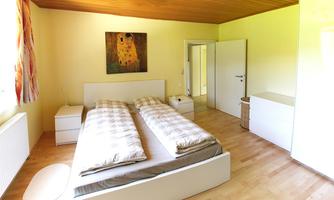 A bright bedroom with a large bed and wooden floor. A painting hangs on the wall, and there are modern furniture.