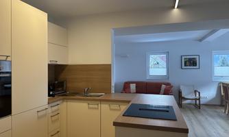 A modern kitchen with light cabinets and wooden details. In the background, there is a cozy living area with a red sofa.