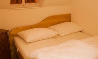 A cozy bedroom with a wooden bed and white pillows. There is a window with bright curtains and a warm wooden floor.