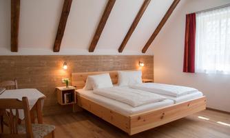 A cozy bedroom with a double bed and a wooden frame. There is a small table, chairs, and large windows for plenty of daylight.