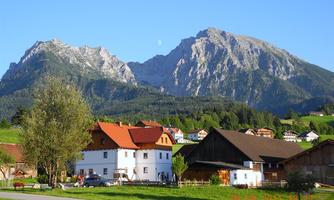 A picturesque landscape with mountains in the background and typical rural houses in the foreground. The sky is clear and blue, creating an idyllic atmosphere.