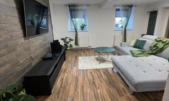 A modern living room with a gray sofa and a flat-screen TV on the wall. The floor is wood, and there are plants as well as a small table in the middle.