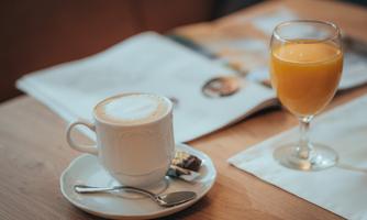 A cup of coffee with milk foam is on a plate next to a glass of orange juice. In the background, a magazine is lying on a wooden table.