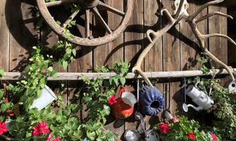A rustic wall with a wooden wheel and various pots. Colorful flowers and plants adorn the lower half of the wall.