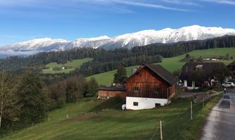 A picturesque landscape with green meadows and snow-capped mountains in the background. In the foreground stands a typical wooden house.
