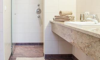 The image shows a modern bathroom with a shower and a sink. On the shelf, there are folded towels and the walls are brightly tiled.