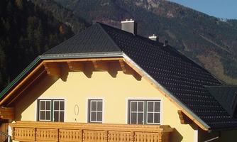 A modern yellow house with a wooden balcony. In the background, majestic mountains and a clear blue sky can be seen.