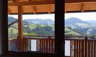 A view from a window onto a green landscape with gentle hills and mountains in the background. The balcony has a wooden railing and invites relaxation.