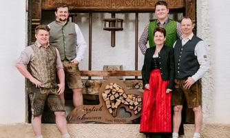 A group of five people stands in front of a historic wooden press. They are wearing traditional Bavarian costumes and smiling warmly at the camera.