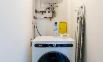 A modern washing machine stands in a narrow laundry room. Behind the machine is a water heater.