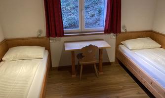 A simple room with two single beds and a small table. Daylight shines in through a window with red curtains.