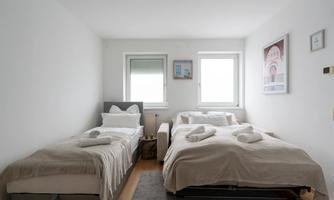 A bright bedroom with two beds and large windows. The walls are painted in neutral colors and decorated with a picture.