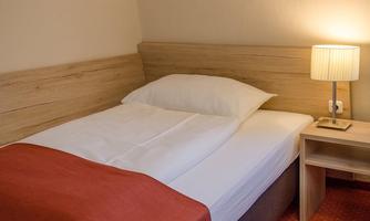 A simple bedroom with a single bed and a modern bedside lamp. The walls are in a light shade, and the bed is decorated with a white duvet and a red throw.