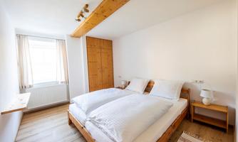 A bright, welcoming bedroom with a double bed and windows with curtains. There is a wooden wardrobe and a small bedside lamp.