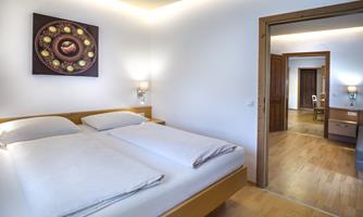 A modern bedroom with two beds and a minimalist design. The room is brightly lit and has an adjacent living room.