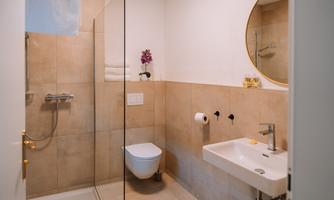 A modern bathroom with a glass shower, a sink, and a wall-mounted toilet. The walls are tiled in beige and there is a round mirror above the sink.