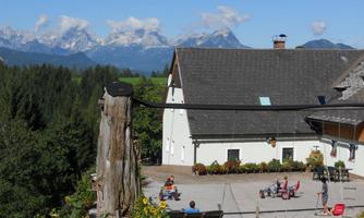 A rural scene with a farmhouse in the foreground and snow-covered mountains in the background. Children are playing on the farm under a clear blue sky.