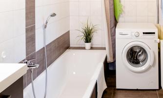 A modern bathroom with a bathtub and a washing machine. The walls are light tiled and there is a green plant on the sink.