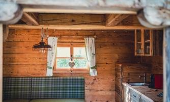 A cozy interior of a wooden cabin featuring a green couch and a well-equipped kitchen. The windows are decorated with white curtains, and there is a lovingly designed atmosphere.