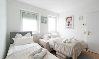 A bright bedroom with two beds and large windows. The walls are painted white and there are some decorations on the walls.