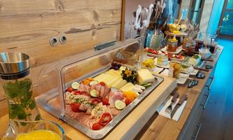 A hearty breakfast buffet with cold cuts, cheese, fresh vegetables, and fruit. Next to it, various drinks and jams are on a wooden tray.
