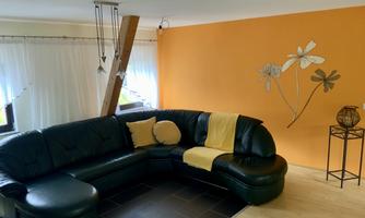 A modern living room with a black couch and an orange accent wall. There are windows with curtains and an interesting wall decoration featuring floral patterns.