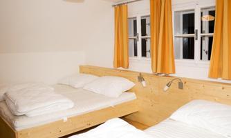 A simple bedroom with two beds and bright orange curtains. The walls are kept neutral, and there are bedside lamps.