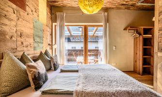 A modern bedroom with bright wood paneling and large windows. The room is cozy furnished with cushions and a soft bed.