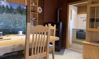A cozy dining area with a wooden table and chairs. In the background, there is a kitchen and a picture of a flower meadow.