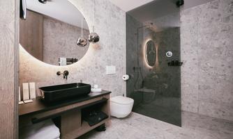 A modern bathroom with an elegant stone wall and large round mirror lighting. The shower is separated by a glass wall, and there is a stylish black sink.