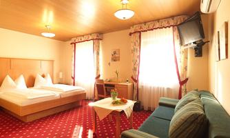 A cozy hotel room with two single beds and a sofa. Bright curtains let in plenty of daylight, and a table with a fresh flower decoration is also visible.