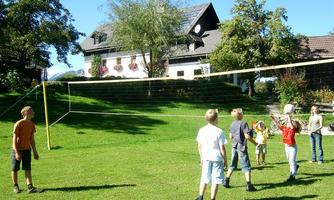 A group of children is playing volleyball on a meadow. In the background, there is a beautiful house with trees and a blue sky.