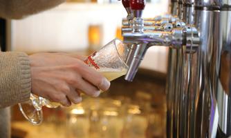 A hand is pouring a glass of beer at a tap. The background features blurred glasses and an inviting atmosphere.