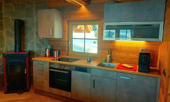 A modern kitchen in a wooden house with a stone wall. It is brightly furnished and has windows that offer a beautiful view.