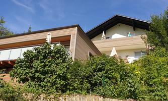 A modern house with two floors and large windows. It is surrounded by green plants and stands under a clear blue sky.