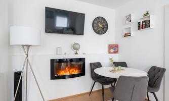 A cozy dining area with a modern table and gray chairs. There is a TV, a clock, and an electric fireplace on the wall.
