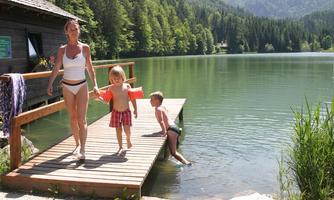 A woman and two children are standing on a dock by the water. In the background, there are forests and mountains visible.
