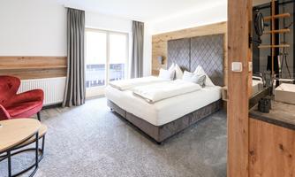 A modern hotel room with a comfortable double bed and a seating area. Large windows bring in plenty of light and offer a view of the balcony.