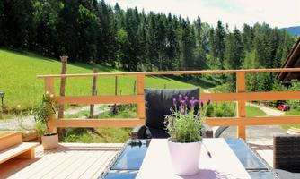 A cozy terrace with a table and a chair, surrounded by green forests. In the foreground is a pot with purple flowers.