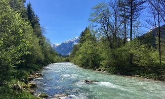 A crystal-clear river flows through a green landscape with trees and mountains in the background. The sky is bright blue and the scene appears tranquil.