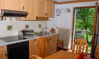 A bright kitchen with wooden cabinets and a table made of light wood. Through the open door, a view of the greenery is visible.