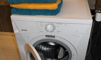 A washing machine stands in a bathroom. On it are stacked towels in blue and yellow, as well as a small space heater.