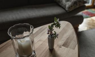 A wooden table with a candle in a glass and a small vase with flowers. In the background, a gray couch is visible.